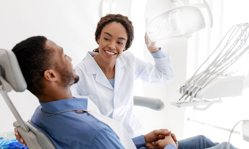 female-dentist-turning-on-lamp-before-treatment,-smiling-to-male-patient Male patient sitting in dental office, female dentist turning on a lamp before treatment, smiling to male patient.